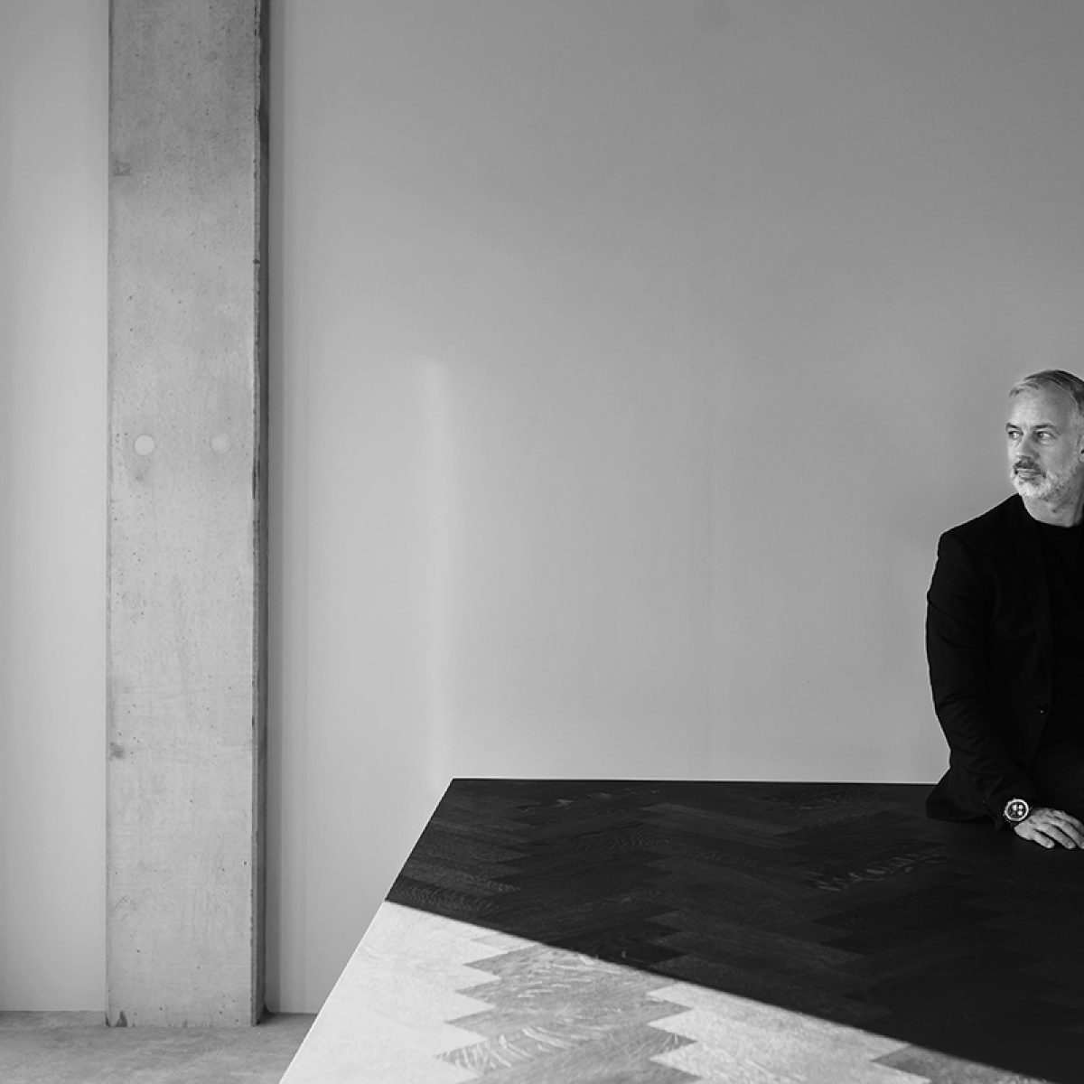 Een omgevingsportret van Arjen van den Hof, gefotografeerd in zijn kantoor met een geconcentreerde, ingetogen sfeer. Het warme, zijlicht accentueert de textuur en diepte van het interieur, terwijl de symmetrische compositie en minimalistische achtergrond verwijzen naar klassieke portrettradities. De verstilde compositie doet denken aan de zorgvuldige omgevingselementen in het werk van Arnold Newman, de grondlegger van de omgevingsportretkunst