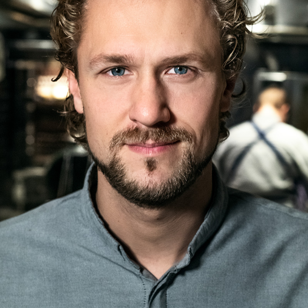 Een omgevingsportret van chef-kok Joris Bijdendijk, vastgelegd in zijn werkruimte met professioneel karakter. De foto combineert gedempte kleurtonen, subtiel contrast en een evenwichtige compositie, waardoor het onderwerp testament wordt van vakmanschap én persoonlijkheid. De benadering draagt verwantschap met de ingetogen intimiteit van Gregory Crewdson en de tijdloze portretten van Annie Leibovitz, waarbij context en sfeer samenkomen in een scherp beeld.
